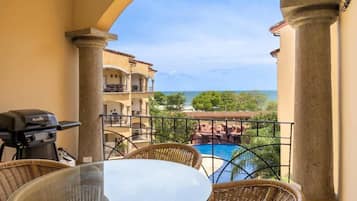 A balcony with a glass table and wicker chairs, overlooking a pool and a view of the ocean and greenery.