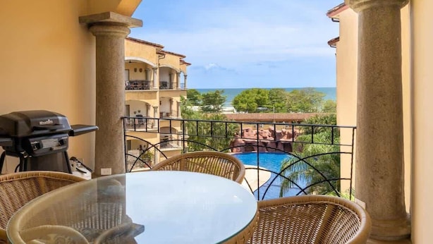 A balcony with a glass table and wicker chairs, overlooking a pool and a view of the ocean and greenery.