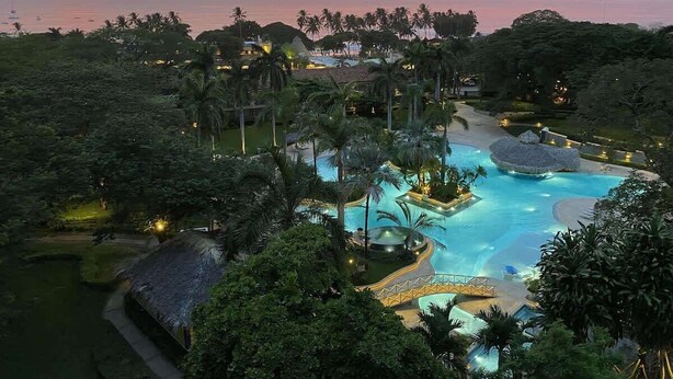 Ein Resort mit einem großen Schwimmbad, umgeben von üppiger Vegetation und Palmen, vor einer Sonnenuntergangslandschaft.