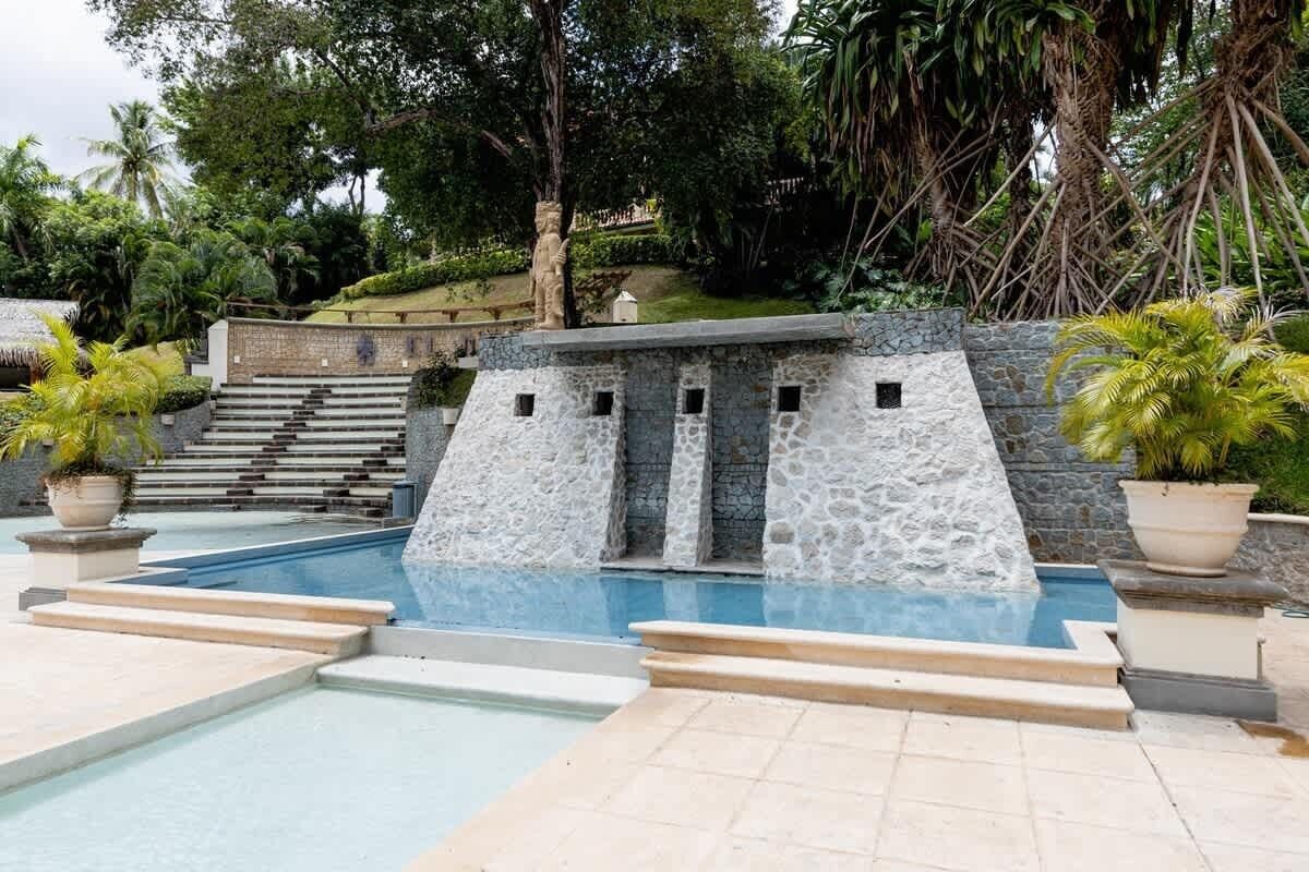 A swimming pool with a stone waterfall feature, surrounded by a stone wall and potted plants.