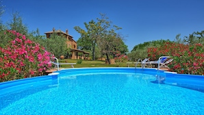 A clear blue swimming pool surrounded by lush greenery and pink flowers, with a stone building in the background.
