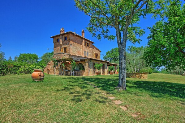 Une maison de campagne en pierre, dotée d’un grand patio et entourée d’une végétation luxuriante, sous un ciel bleu azur.