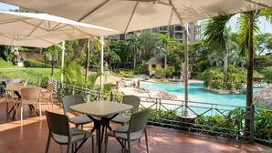 A covered outdoor seating area with tables and chairs, overlooking a swimming pool and lush greenery.