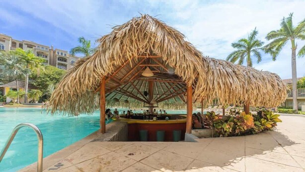 A poolside bar with a thatched roof, surrounded by palm trees and a pool.