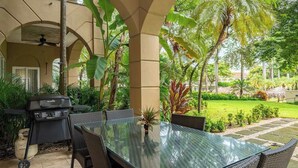 An outdoor patio with a glass-top table and chairs, surrounded by lush greenery and a view of a manicured lawn. - Matapalo 102- Poolside Condo at the Diria Resort- (Tamarindo)