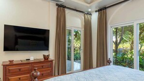 A bedroom with a large bed, a flat-screen TV mounted on the wall, a wooden dresser, and a sliding glass door leading to a balcony with a view of greenery. - Matapalo 102- Poolside Condo at the Diria Resort- (Tamarindo)
