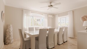Un coin repas avec une table et des chaises blanches, un ventilateur de plafond et de grandes fenêtres ornées de rideaux légers.