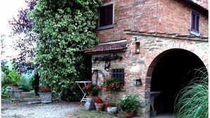 Un edificio rustico in pietra con un'ampia entrata ad arco, circondato da un giardino con vasi di piante e fiori.