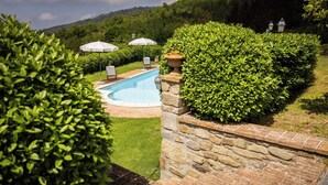 Una piscina rodeada de exuberante vegetación, muros de piedra y mobiliario exterior con sombrillas.