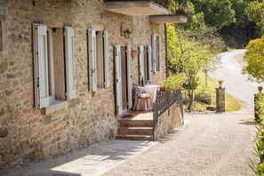 A stone building with multiple windows, a table with chairs, and a gravel path leading to a road.