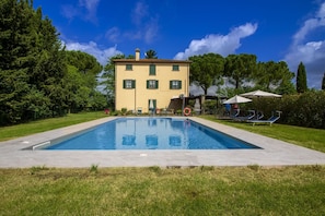 Ein rechteckiger Pool mit einer gefliesten Kante, ein Gebäude mit gelber Fassade und grünen Fensterläden sowie ein klarer, blauer Himmel.