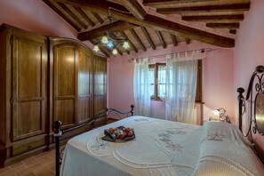 A bedroom with wooden wardrobe, a bed with a white bedspread, a hanging light fixture, and a window with sheer curtains.
