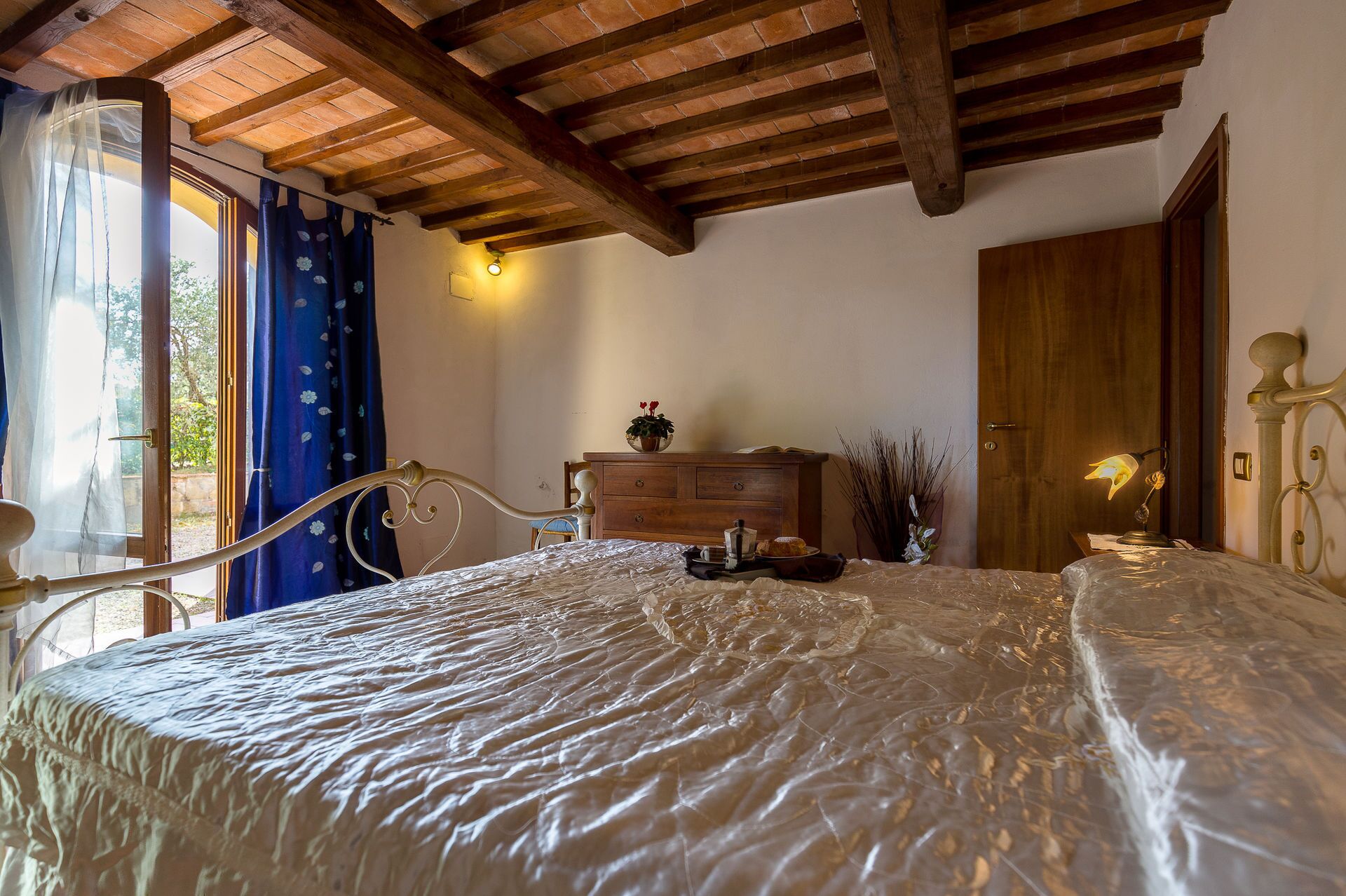 A bedroom with a wooden ceiling, a bed with a white bedspread, a wooden dresser, a nightstand with a lamp, and a window with curtains.