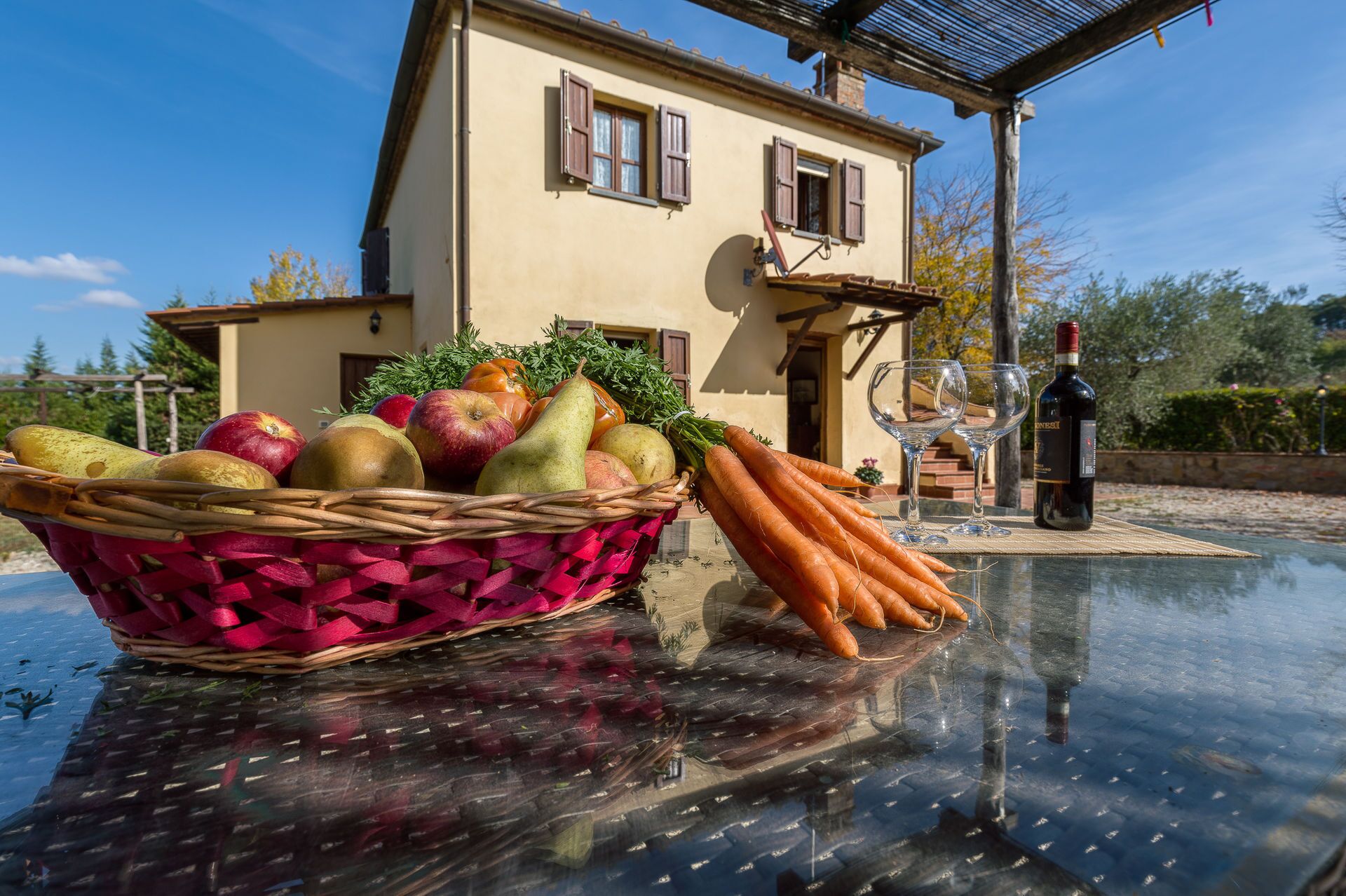 A house with a large basket of fruits and vegetables on a glass table, a bottle of wine, and a clear sky.