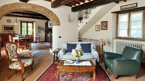 A spacious living room with a high ceiling, exposed wooden beams, a large archway, a sofa, a green armchair, and a dining area in the background.