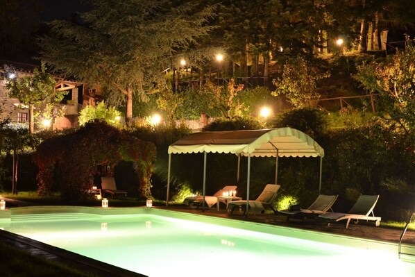 Uma área de piscina à noite, com espreguiçadeiras e um espaço coberto para sentar, cercada por árvores e um edifício ao fundo.