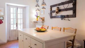 A kitchen with a central island, pendant lights, a wooden shelf with decorative items, and a door leading outside.