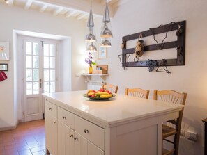 A kitchen with a central island, pendant lights, a wooden shelf with decorative items, and a door leading outside.
