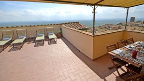 An outdoor terrace with sun loungers, a table set for dining, and a view of the sea and distant buildings. - Beautiful apartment for 4 people with A/C, WIFI, TV, terrace, pets allowed and panoramic view (Cortona)