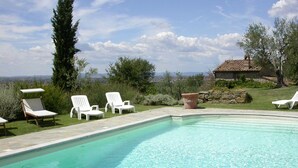 Une piscine à l’eau claire, entourée de chaises longues blanches, avec une maison en pierre en arrière-plan.