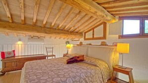 A cozy bedroom with a wooden ceiling, a large bed with patterned bedding, a bedside table with a lamp, and a window providing natural light. - Capannino - Holiday House Rental near Siena, Tuscany (Ponte A Bozzone)