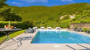 A swimming pool with lounge chairs and a table with fruit and a bottle, surrounded by a lush green hillside.