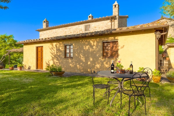 A stone building with a tiled roof, a wooden door, and a window. There is a table with chairs and a bottle of wine.