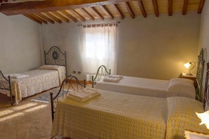 A bedroom with two single beds, a window with curtains, a table with a tablecloth, and a lamp on a bedside table.