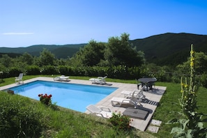 Ein rechteckiger Pool mit Liegestühlen und einem Tisch, umgeben von viel Grün und Hügeln im Hintergrund.