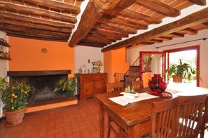 A rustic interior with exposed wooden beams, a fireplace, a dining table with chairs, and potted plants.
