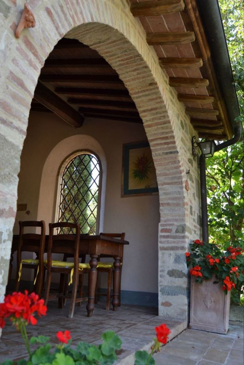 An arched stone entrance with a wooden table and chairs, a window with a lattice design, and a potted plant with red flowers.