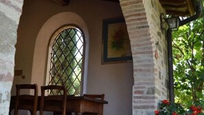 Une entrée en pierre en forme d’arche, avec une table et des chaises en bois, une fenêtre ornée d’un motif en treillis et un pot de fleurs contenant des fleurs rouges.