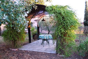 A covered outdoor seating area with a table and chairs, surrounded by greenery and a pergola structure.