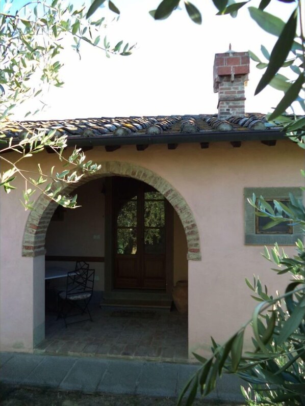 A building with a tiled roof, a brick chimney, and an arched doorway leading to an interior space with a chair and a table.