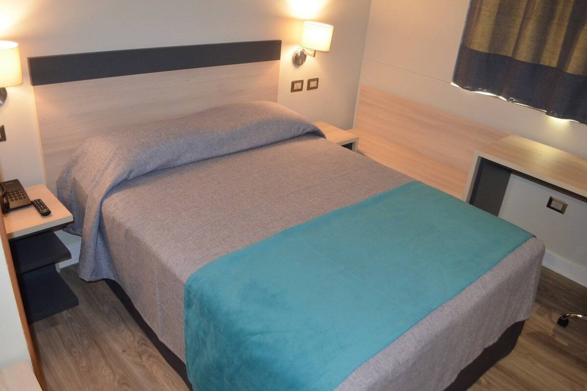 Una cama individual con colcha gris y azul, una mesita de noche con teléfono y una lámpara fijada a la pared.