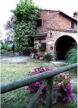 A rustic stone building with a large arched entrance, surrounded by a garden with potted plants and flowers.