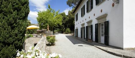 A cobblestone street leading to a white building with black shutters, surrounded by greenery and a stone wall.