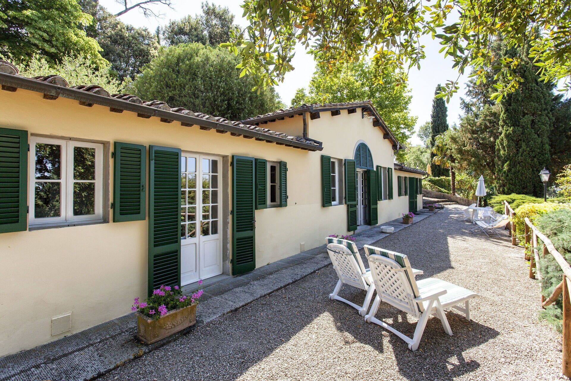 A building with a tiled roof, green shutters, and a gravel driveway. There are outdoor seating areas with white chairs and a table.