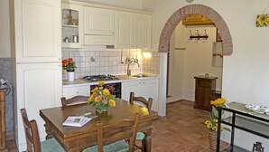A kitchen with white cabinets, a stove, and a sink. There is a dining table with chairs and a vase of flowers. An archway leads to another room. - Stunning private villa for 4 people with WIFI, private pool, TV, terrace and panoramic view (Cortona)