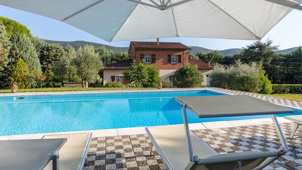 A swimming pool with lounge chairs and an umbrella, surrounded by a house and greenery.