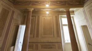Un salón imponente con techo y columnas ornamentadas, un elemento decorativo circular grande y una puerta que conduce a otra habitación.