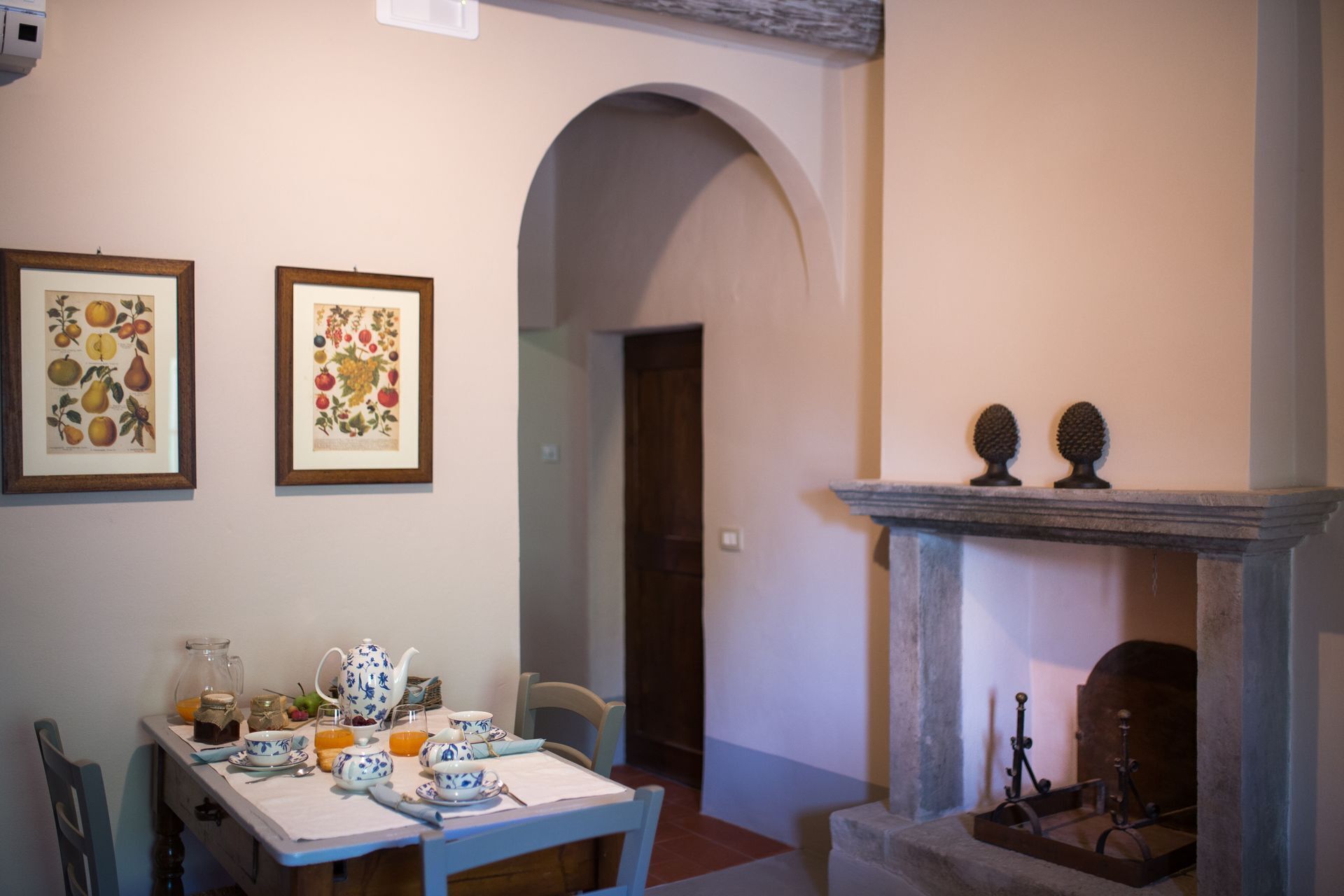 A dining area with a table set for breakfast, a fireplace with two decorative urns, and two framed botanical prints on the wall.