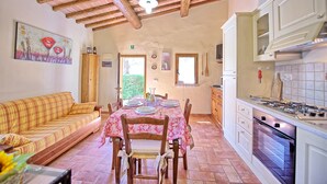 Un coin repas accueillant, avec une table en bois dressée pour quatre personnes, entourée de chaises en bois. Un canapé est placé au premier plan, et une cuisine avec des armoires blanches et un four est visible sur la droite.