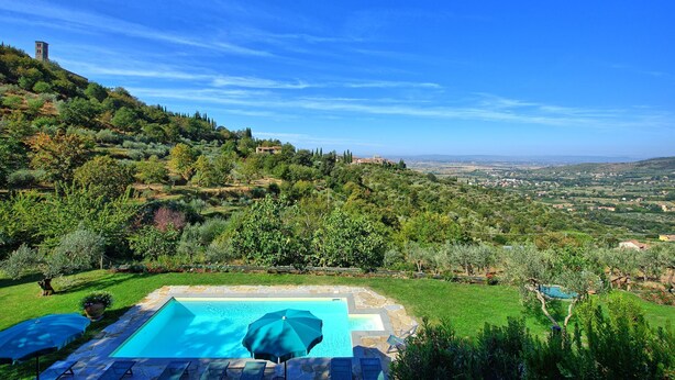 A swimming pool with lounge chairs and umbrellas, surrounded by lush greenery and a view of a distant town.