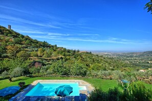 A swimming pool with lounge chairs and umbrellas, surrounded by lush greenery and a view of a distant town.