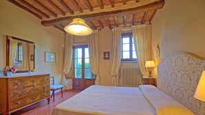 A bedroom with a wooden dresser, a bed with a white bedspread, a nightstand with a lamp, and a window with curtains.