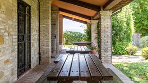 Una veranda coperta con tavolo e panche in legno, pareti in pietra e vista sul verde.