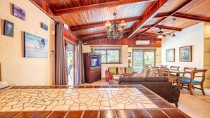 A spacious living area with a large wooden dining table, a sofa, and a TV. There is a ceiling fan and a chandelier.