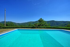 A rectangular swimming pool with clear blue water, surrounded by a hedge and trees, with a mountain range in the background.