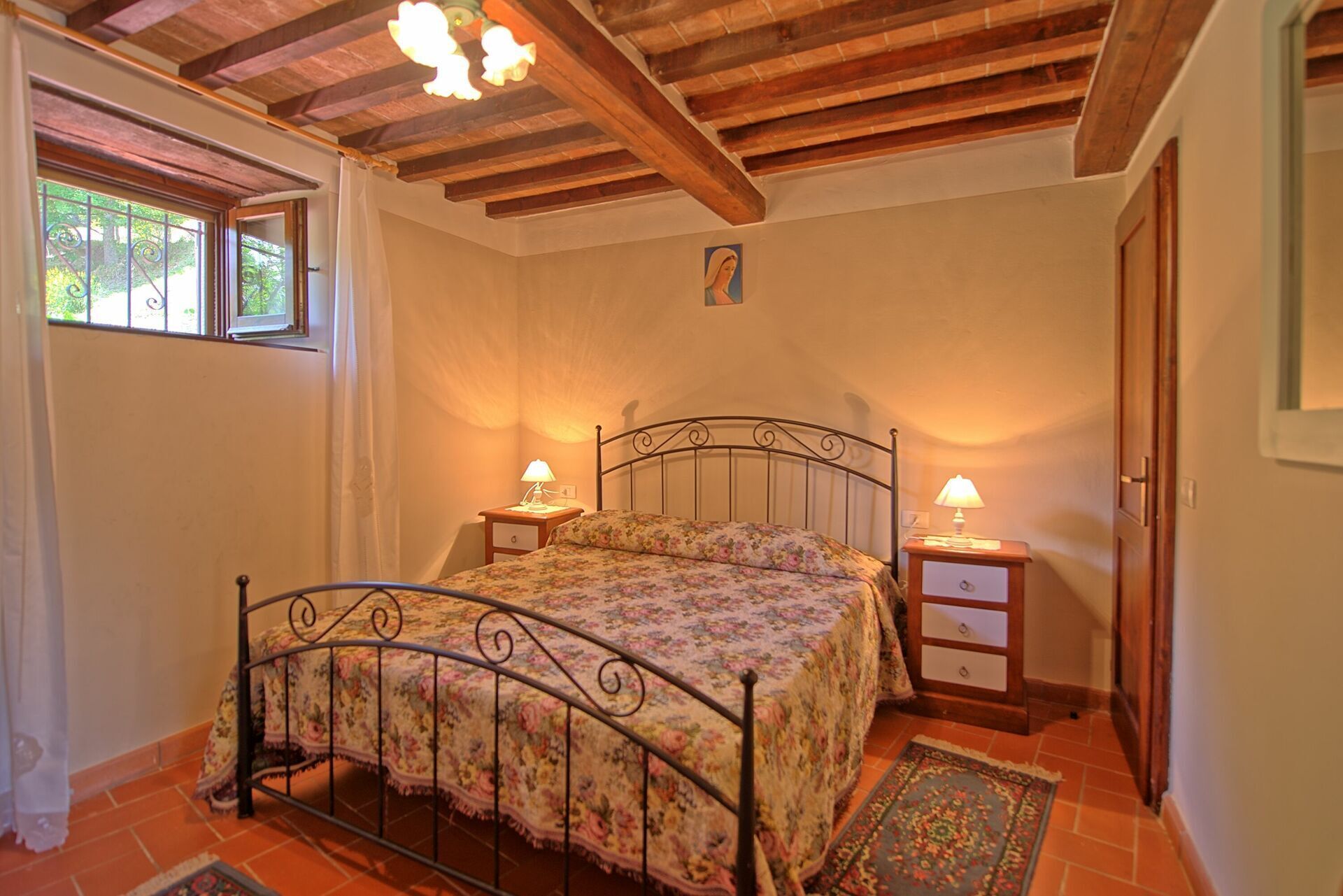 A bedroom with a bed, two bedside tables, a nightstand, a wooden ceiling, a window with bars, and a door.